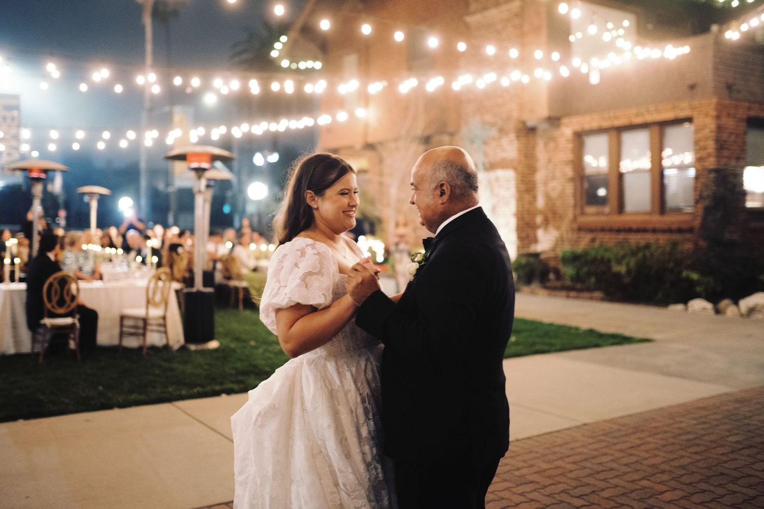 Bride dances with older gentleman under warm string lights at elegant outdoor evening wedding.