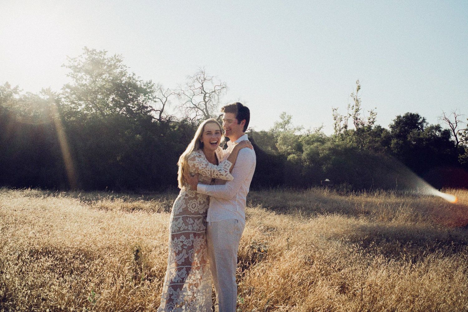 Couple embracing in sunlit golden field with trees in background, woman in floral dress and man in light suit.
