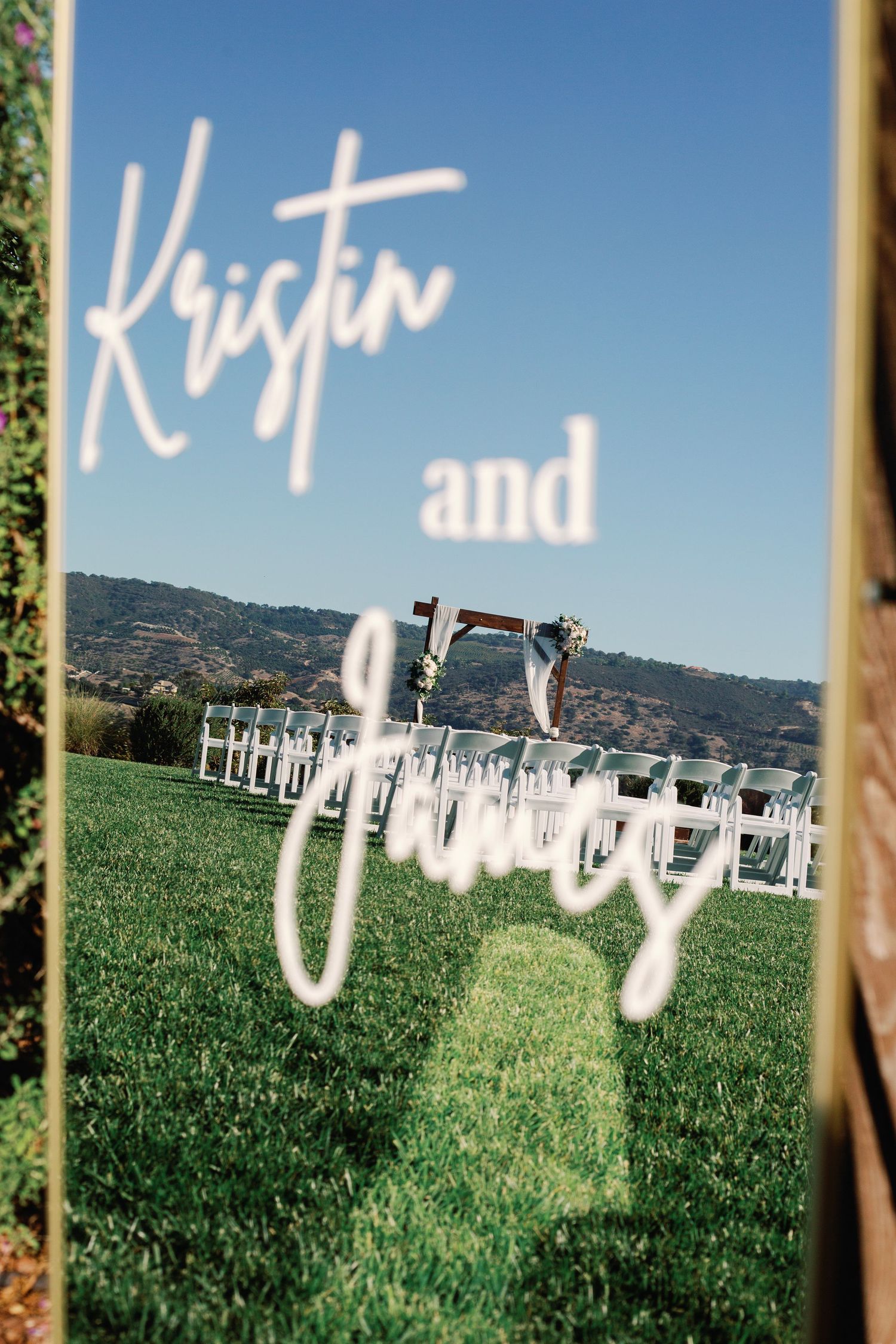 Wedding ceremony reflected in a mirror sign reading Kristin and James with outdoor altar and mountain backdrop.