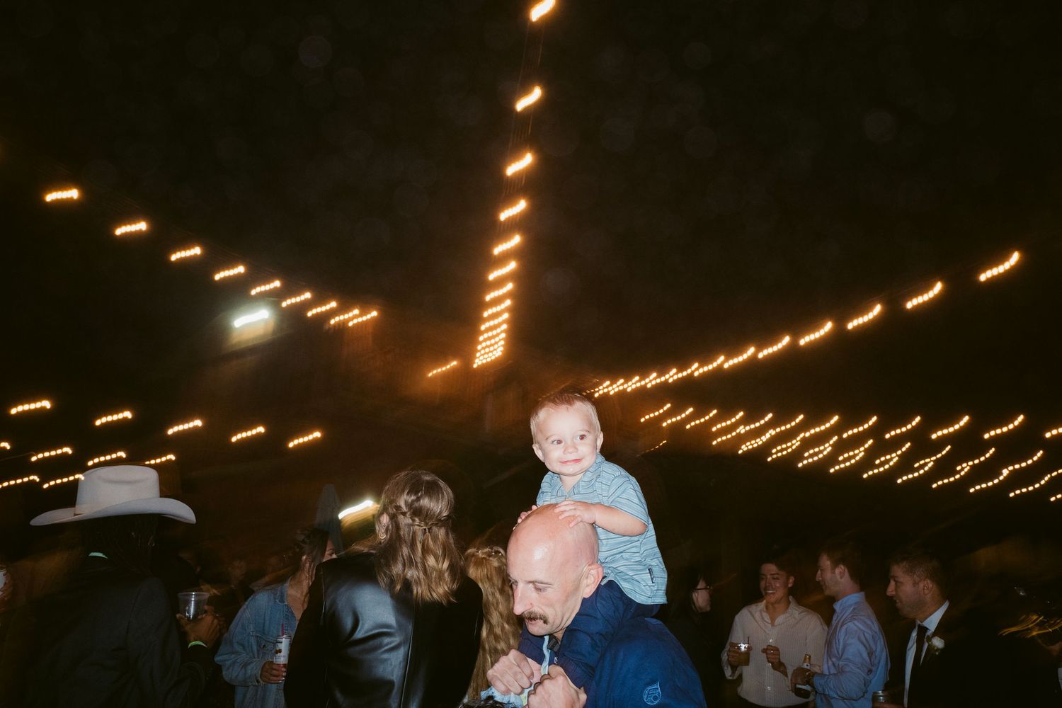 Baby sitting on mans shoulders at lively outdoor wedding reception under string lights at night.