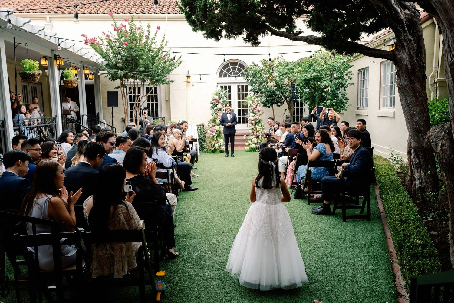 Flower girl walks down the aisle at an outdoor garden wedding ceremony with seated guests.