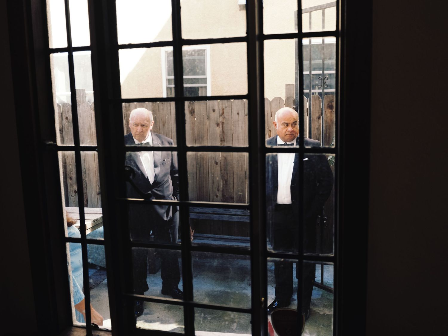Two elderly men in dark suits viewed through French doors with iron grid panes at a wedding venue.