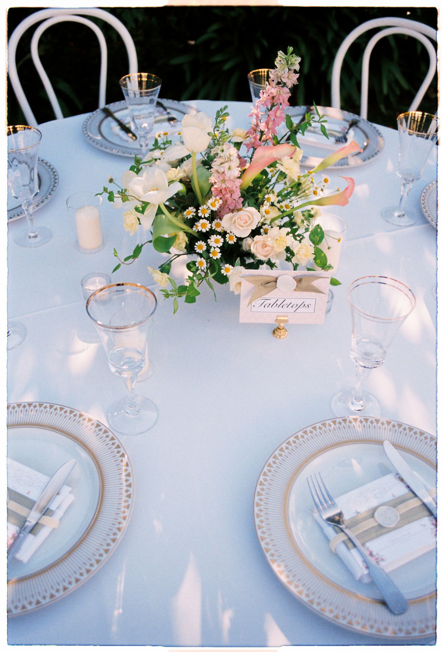 Round wedding reception table with gold-rimmed charger plates, crystal glassware, and a pink and white floral centerpiece.