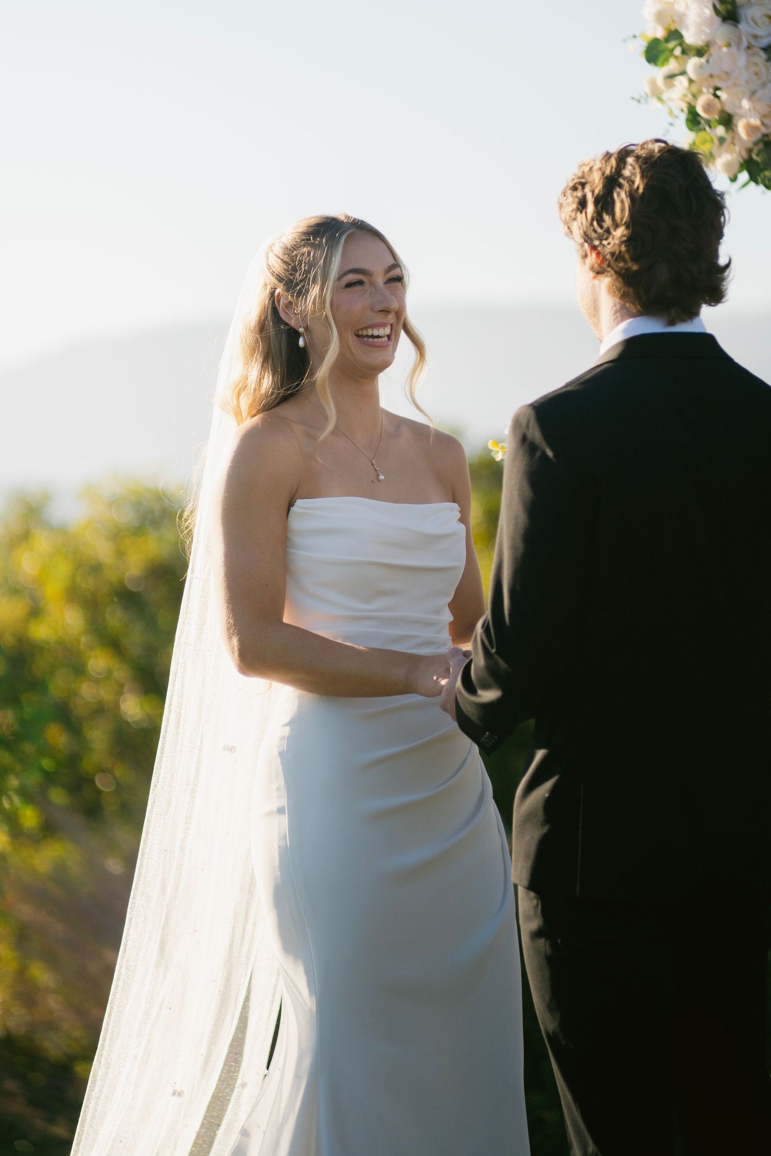 Radiant bride in strapless white gown and veil smiling at groom during outdoor wedding ceremony.