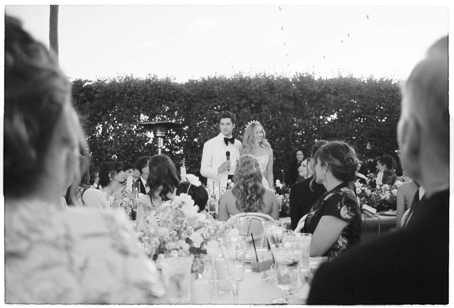 Black and white outdoor wedding reception with couple giving speech at elegantly decorated table surrounded by guests.