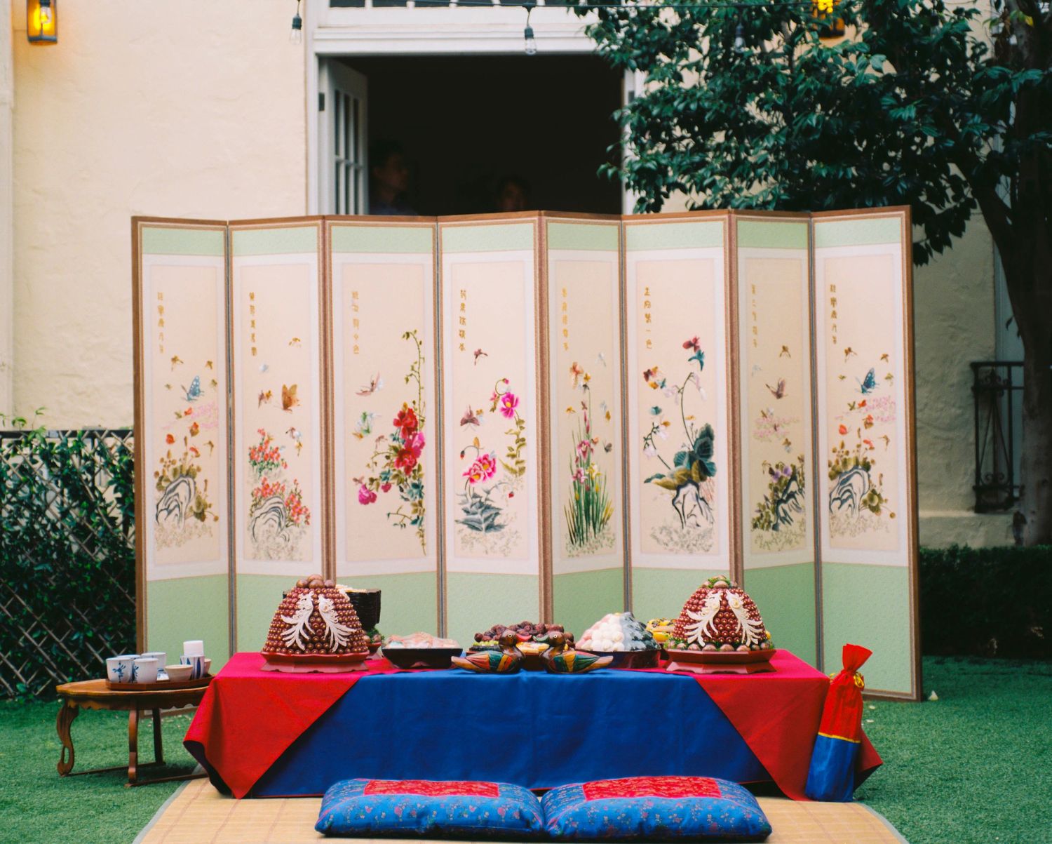 Colorful floral folding screen backdrop with low table set for traditional Korean wedding ceremony outdoors.