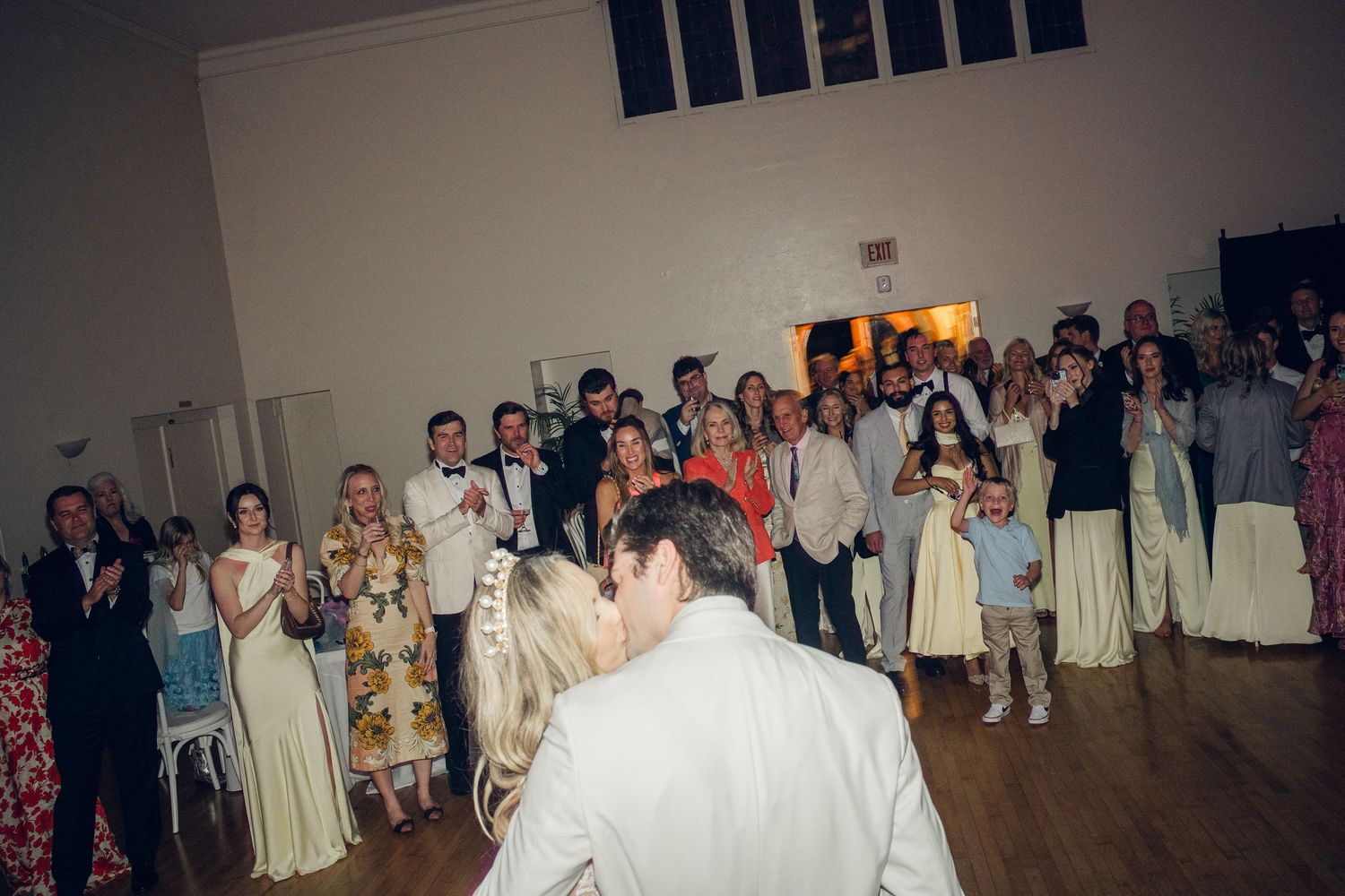 Bride and groom share their first dance as wedding guests clap and watch in a bright reception hall.