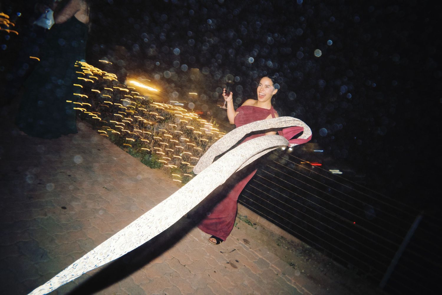 Woman in elegant burgundy gown posing near a long white ribbon at a nighttime wedding reception.