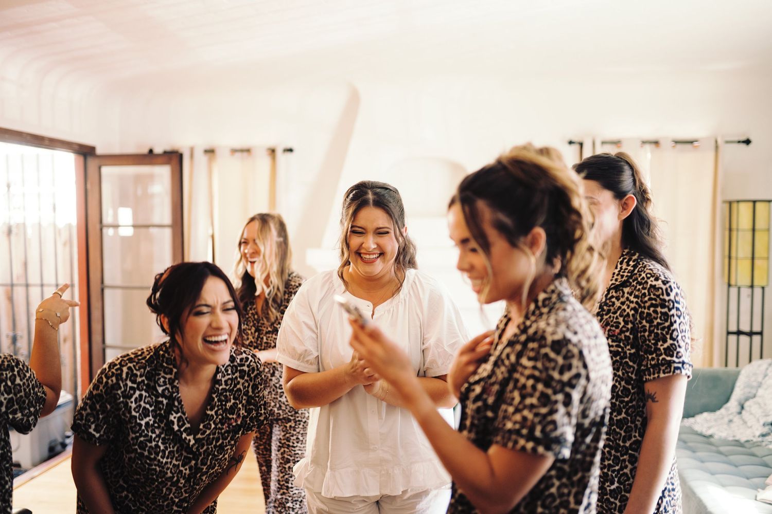 Bride laughing joyfully with bridesmaids in leopard print robes during fun wedding morning preparations.