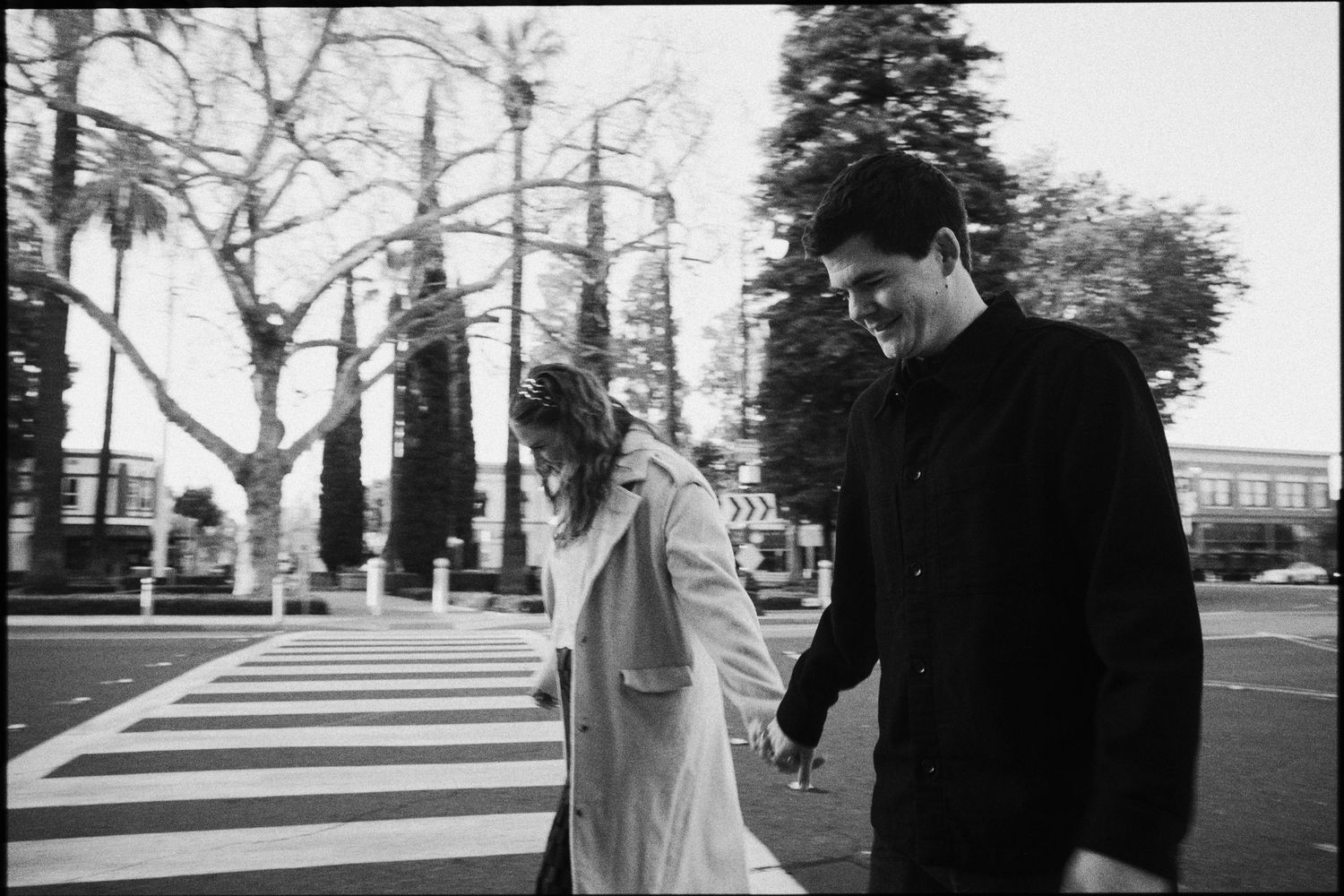 Black and white photo of a smiling couple holding hands while crossing a street in a tree-lined urban setting.
