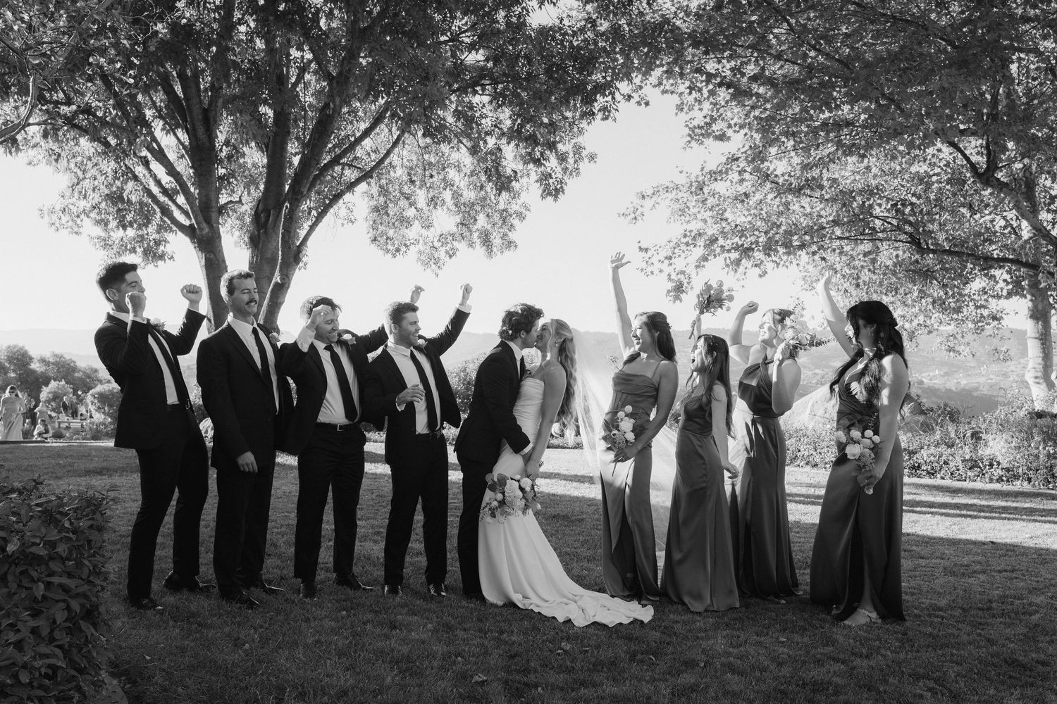 Black and white photo of wedding party celebrating under trees with bridesmaids and groomsmen cheering.