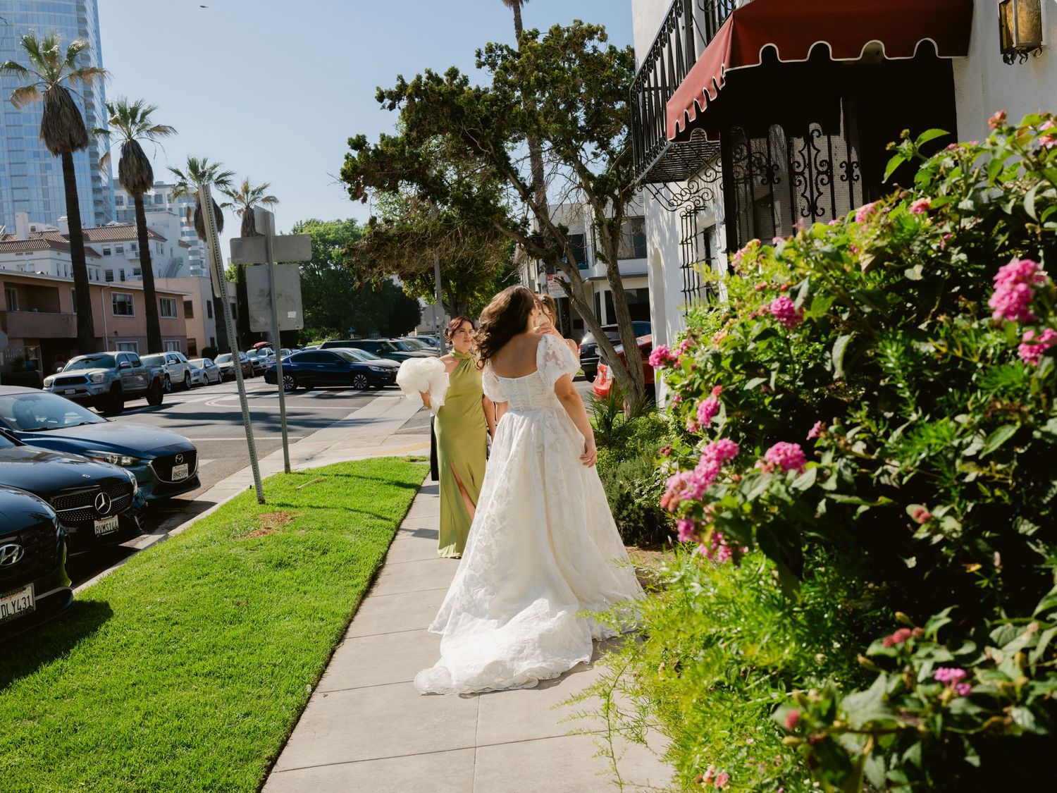 Bride in white ball gown walks along a sunny urban sidewalk with pink roses and palm trees nearby.