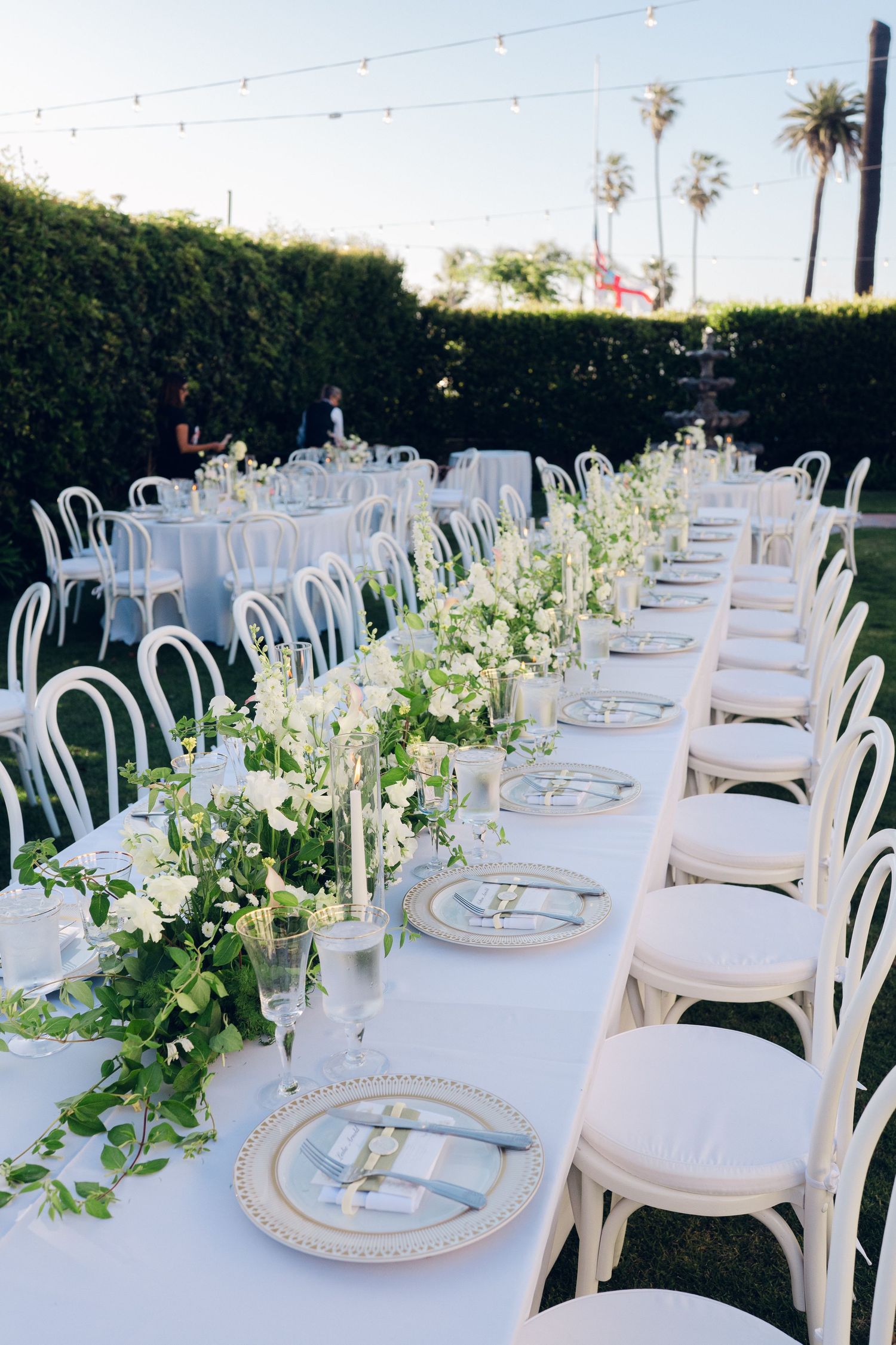 Elegant outdoor wedding reception with long white tables, greenery centerpieces, and white chairs.