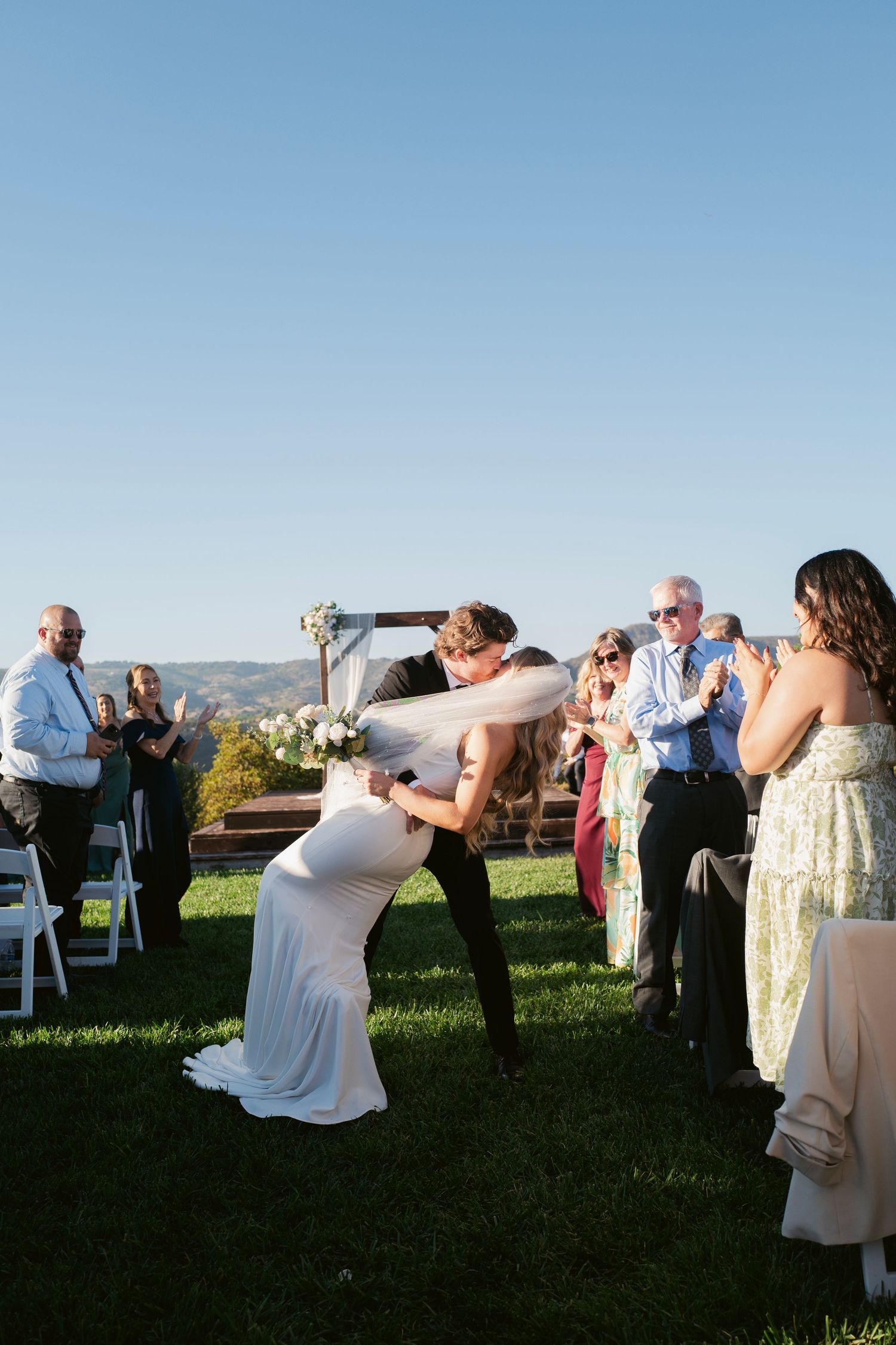 Groom dipping bride in white gown for romantic kiss while guests cheer after outdoor ceremony.
