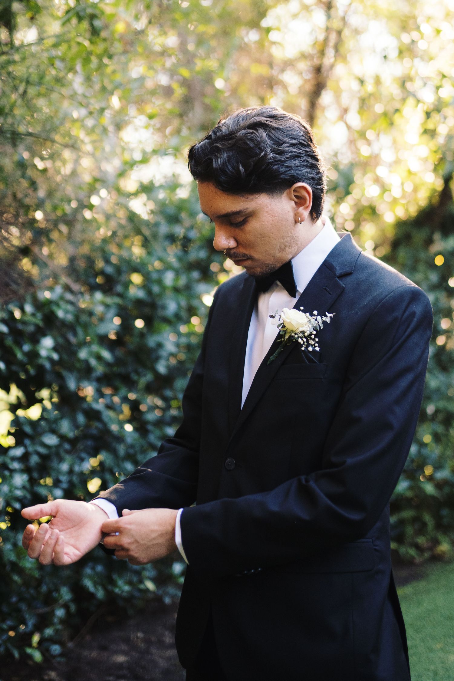 Groom in black tuxedo with white boutonniere adjusting cufflinks outdoors.
