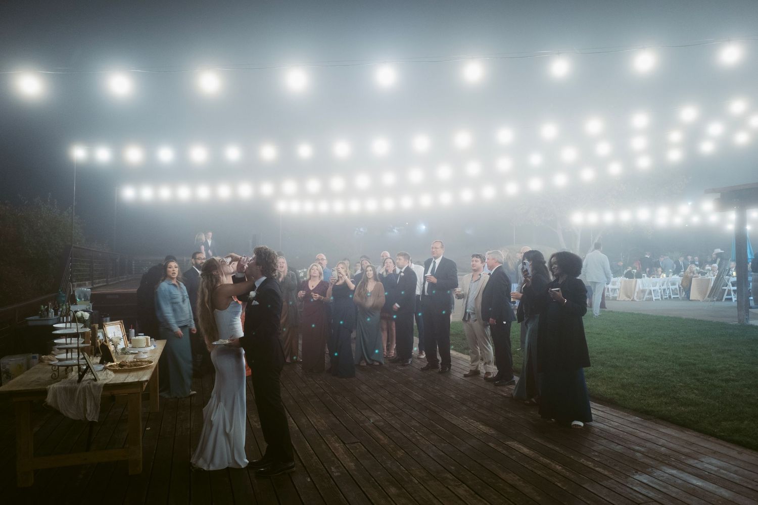 Wedding guests socializing on a wooden deck under glowing string lights at night.