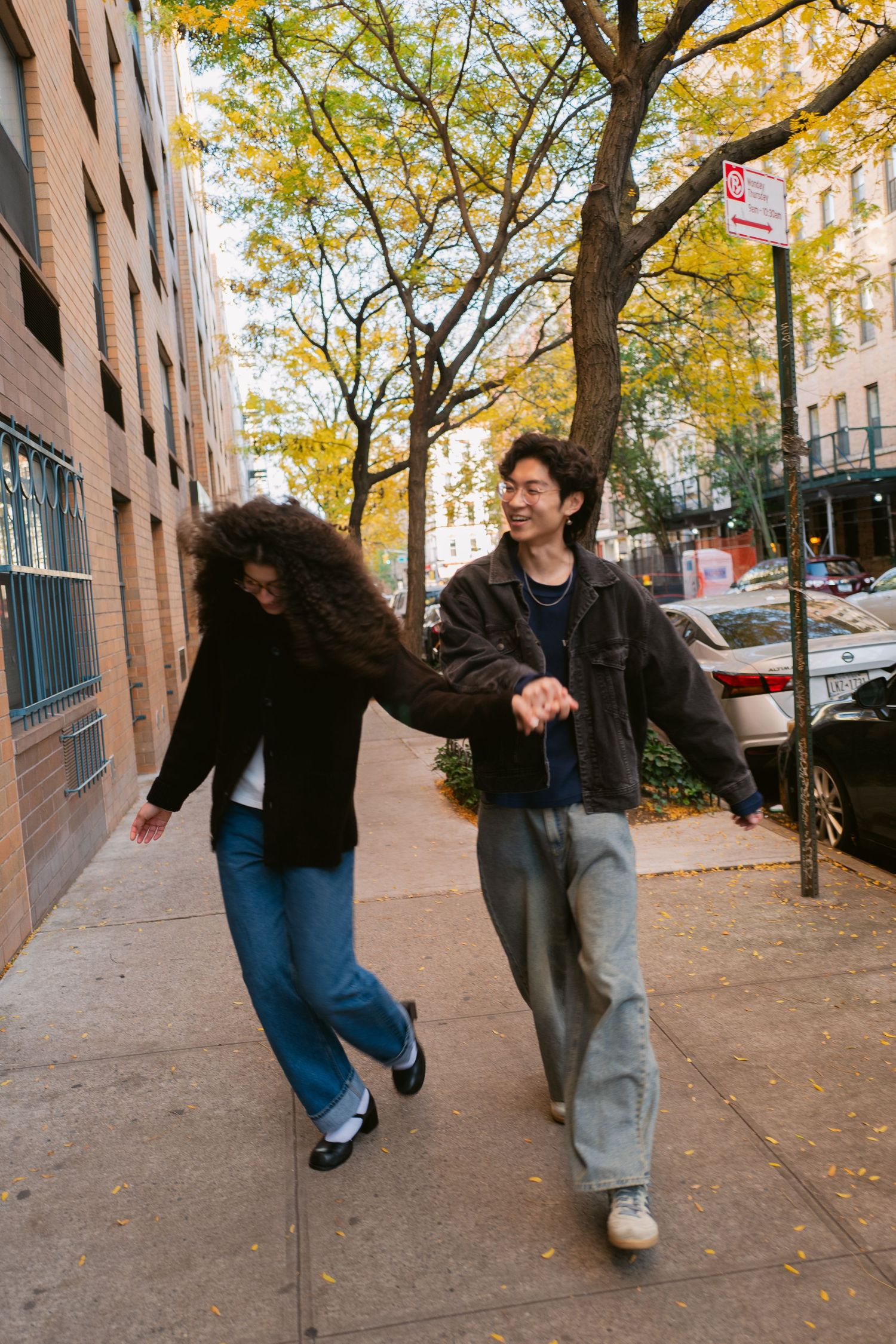 A joyful couple laughs and holds hands while strolling along an urban sidewalk with autumn trees.