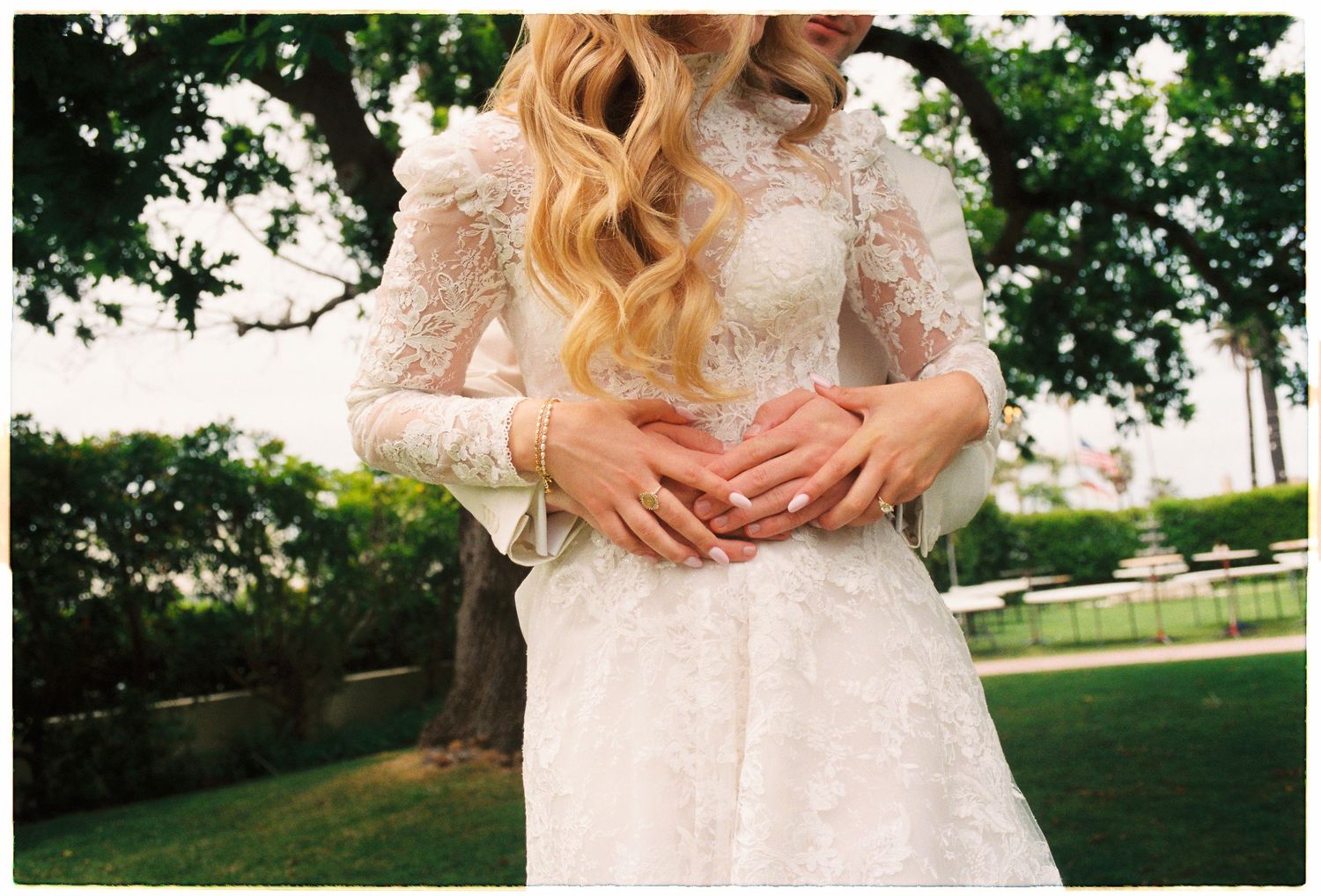 Bride in long-sleeve lace wedding dress with flowing blonde hair stands outdoors with hands clasped together.
