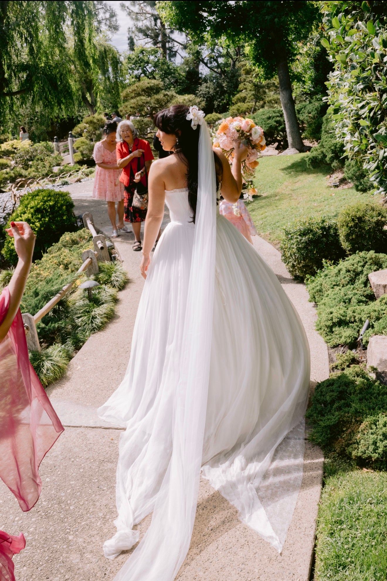 Bride in white strapless gown with long veil and floral crown walks garden path holding bouquet.