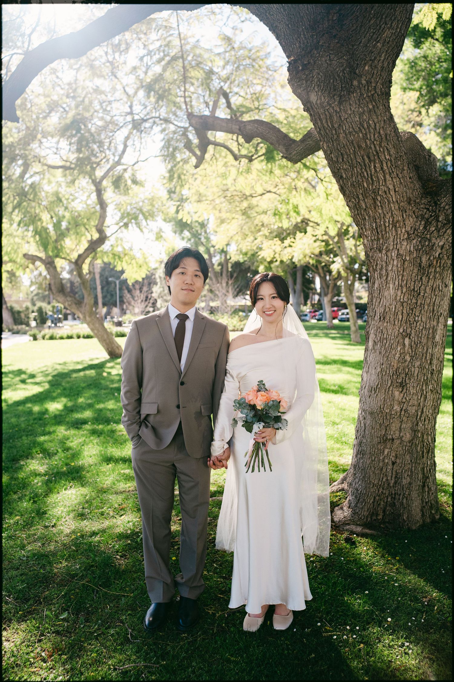 A couple in wedding attire poses under a large tree in a sunlit park, the bride holding a peach floral bouquet.