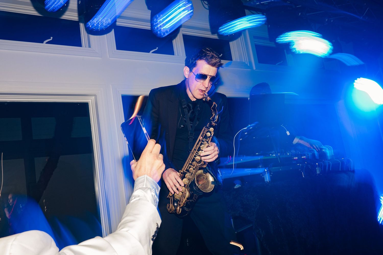 Saxophonist in black suit and sunglasses performs under blue stage lights at a lively indoor event.