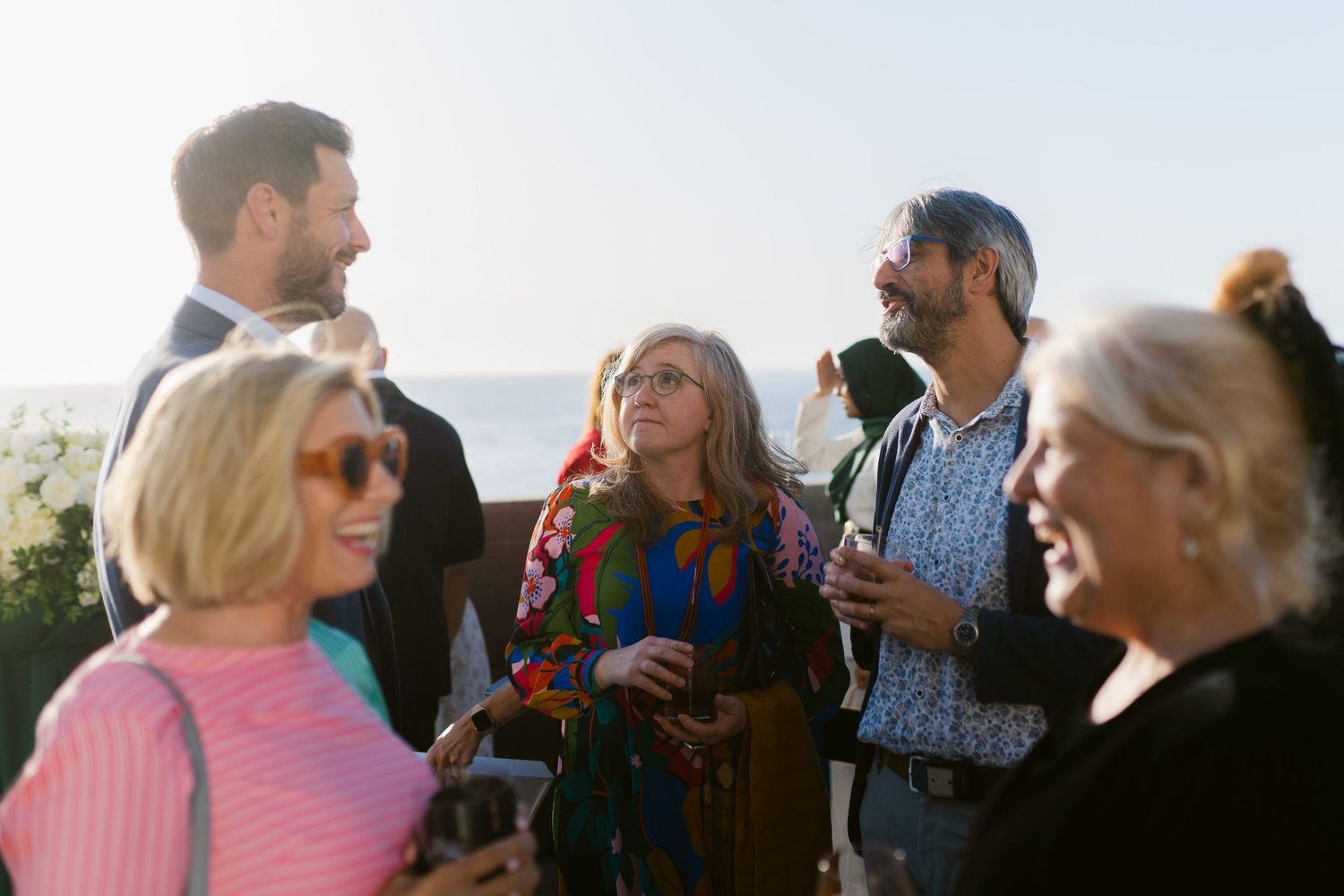 Group of adults socializing outdoors at a sunny gathering, holding drinks and enjoying conversation against a scenic backdrop.