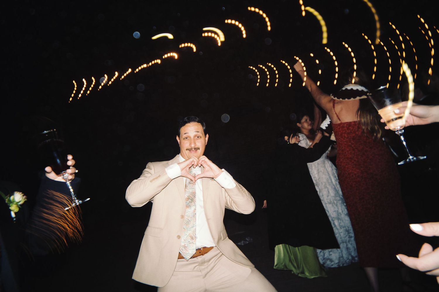 Man in beige suit making a heart gesture while dancing at a festive wedding reception.
