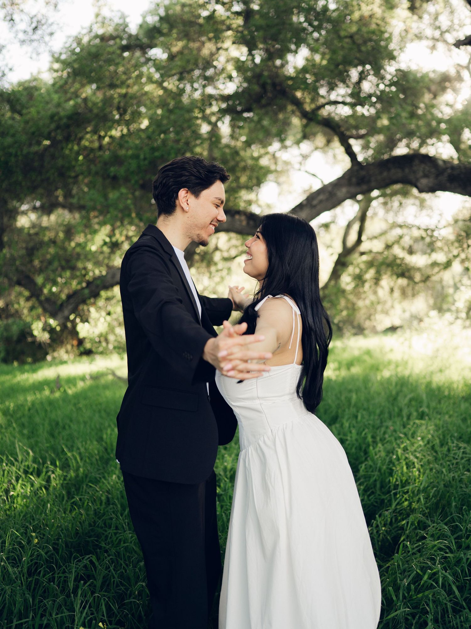 Engaged couple gazes lovingly at each other while holding hands in sunlit green field.