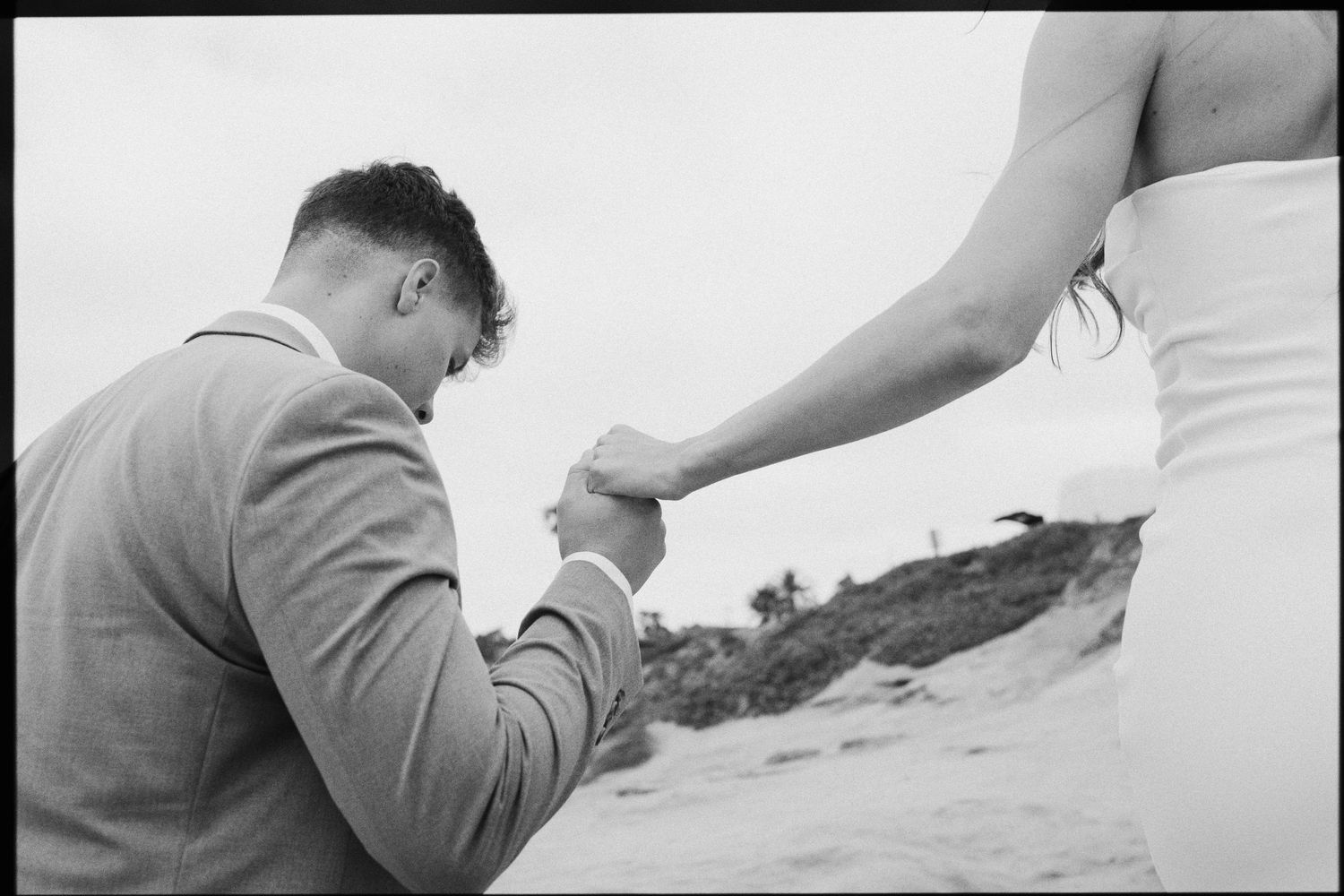 Black and white photo of a man kneeling to hold his partners hand on the beach.