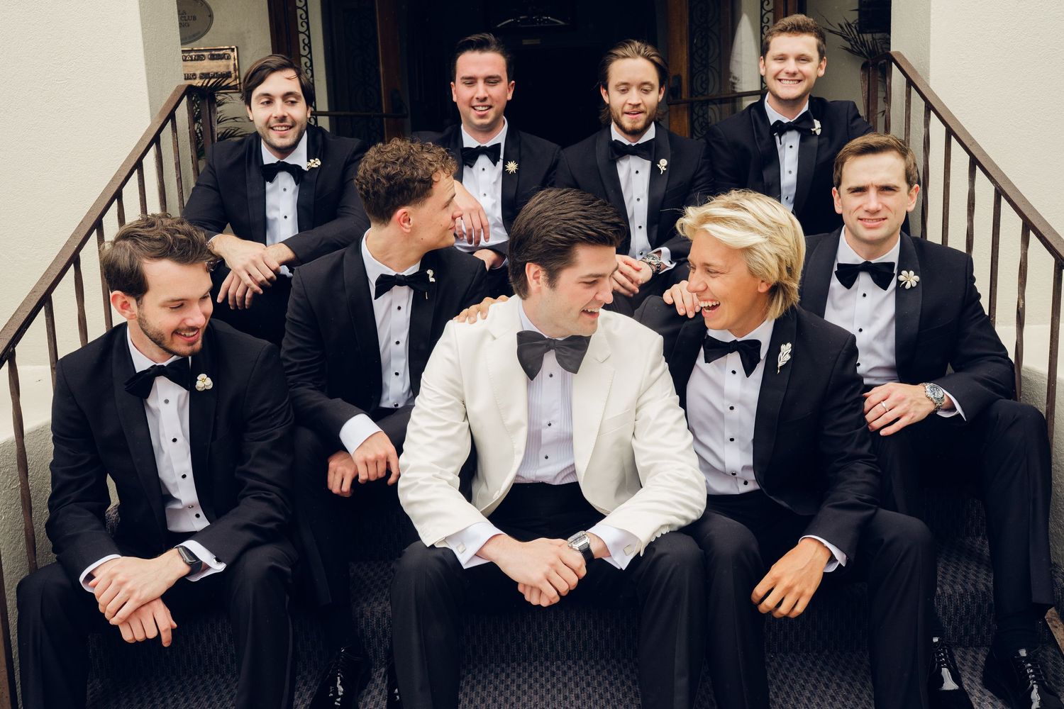 Group of nine groomsmen in black and white tuxedos with bow ties posing together on staircase steps.