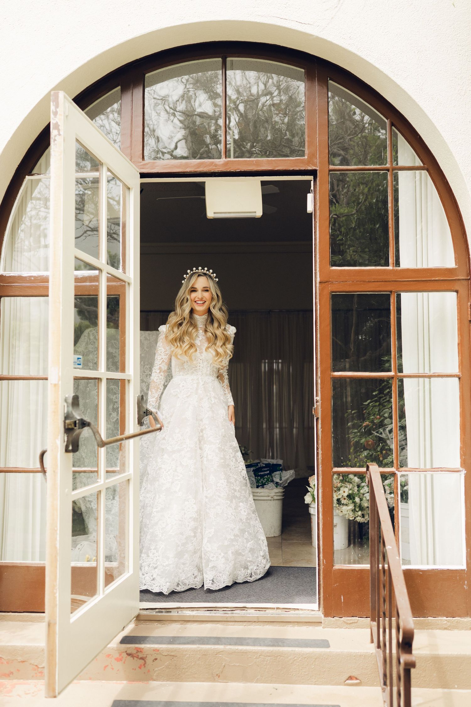 Elegant bride in lace long-sleeve wedding gown and crystal crown standing in arched French doors.
