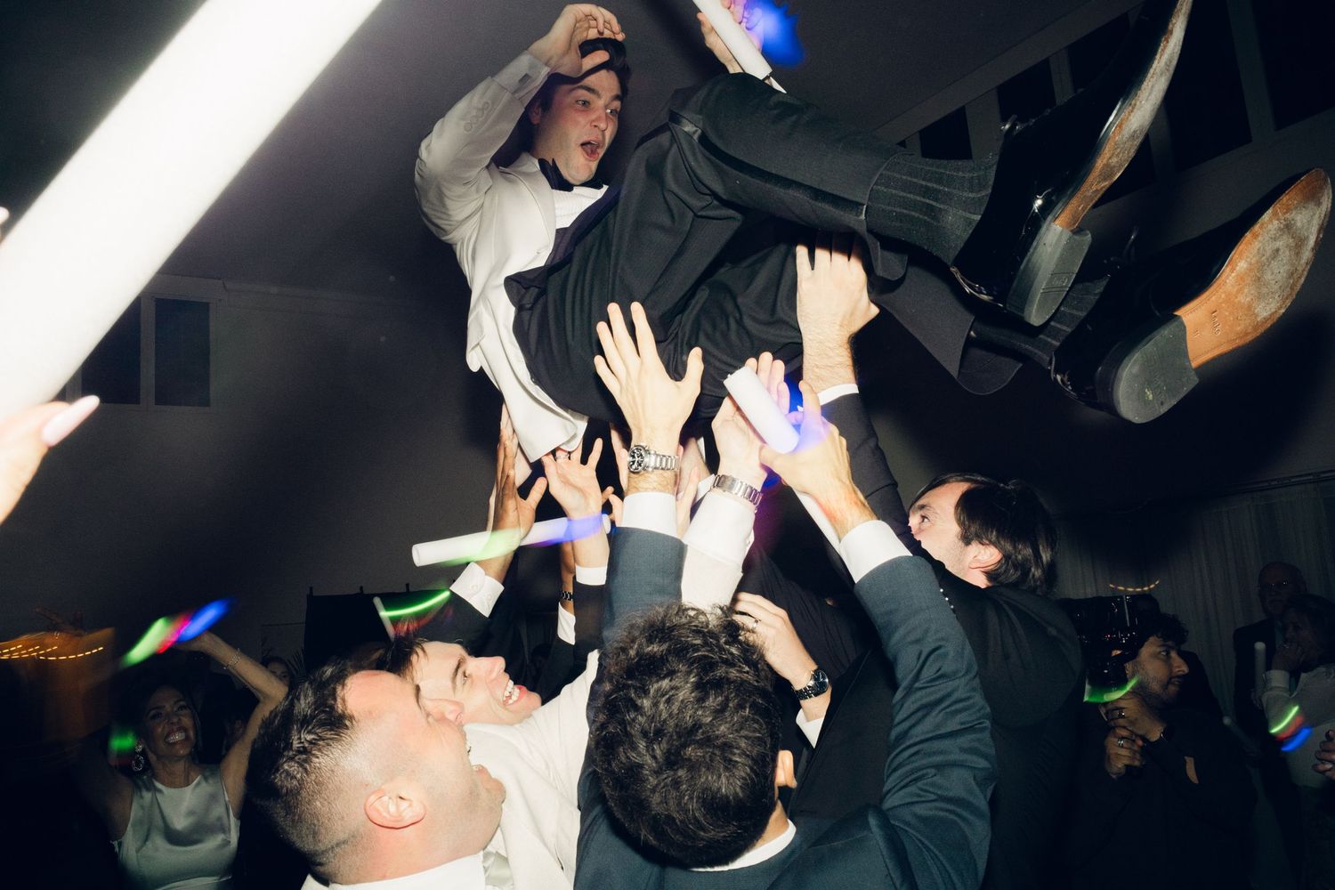 Groomsmen lift the groom above the crowd during a lively wedding reception dance floor celebration.
