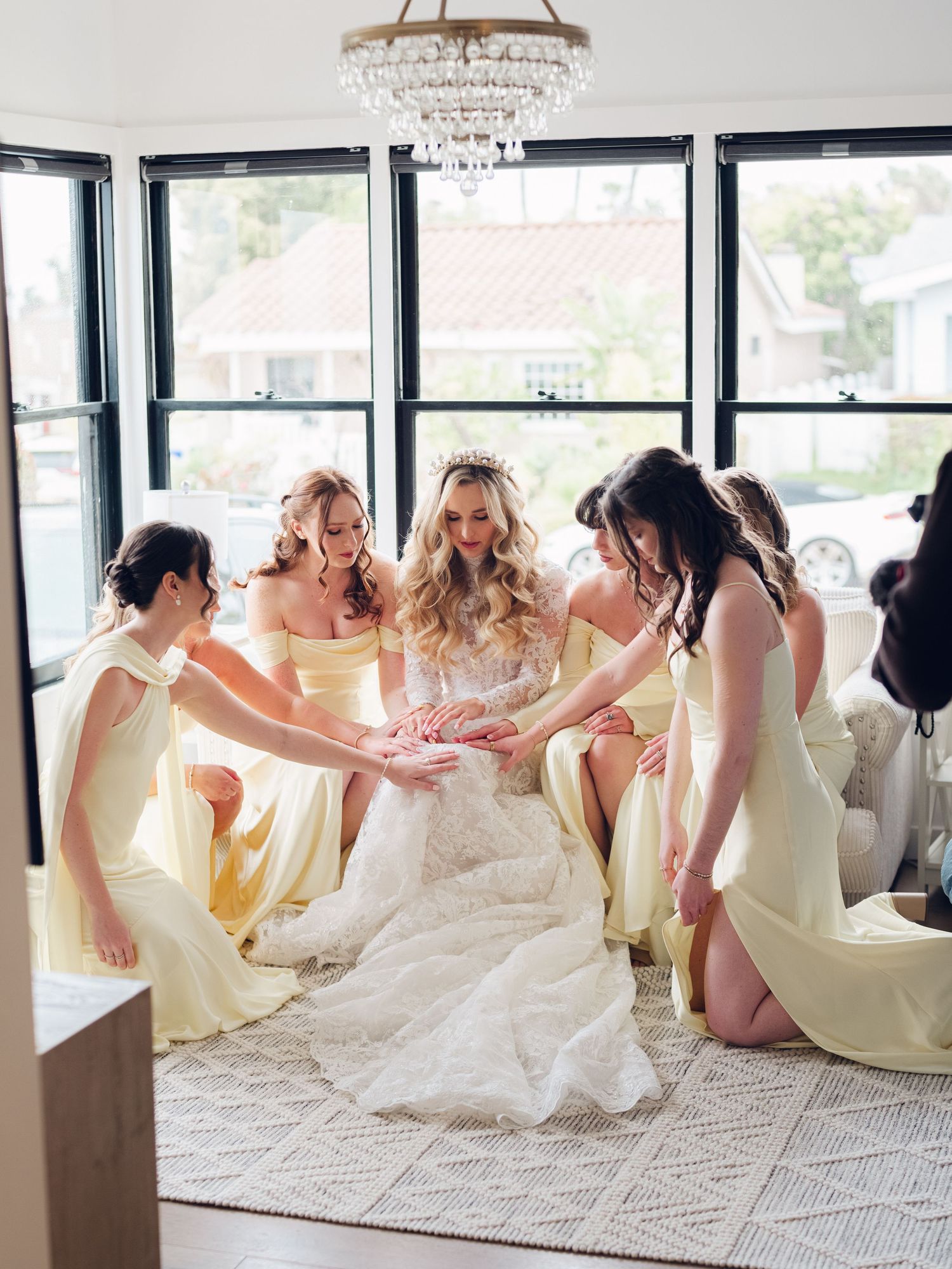 Bride in lace wedding gown surrounded by bridesmaids in yellow dresses reaching toward her hands in a bright indoor setting.