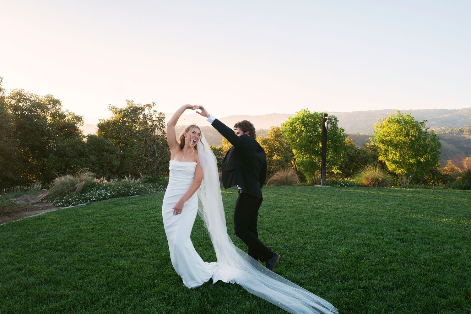 Groom twirling bride in white gown and cathedral veil on a green lawn at golden hour sunset.