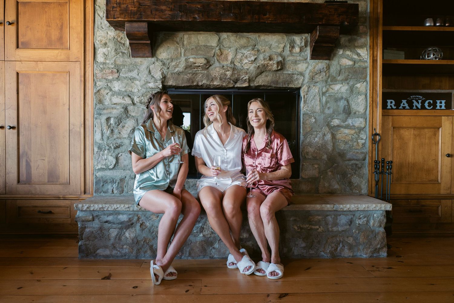 Three bridesmaids in colorful satin pajamas sitting and laughing by a rustic stone fireplace.