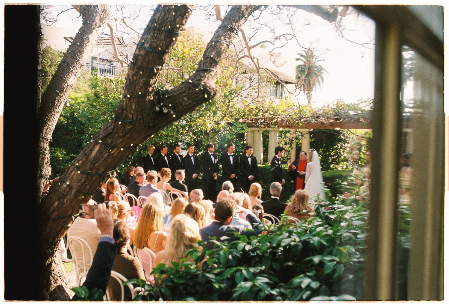 Outdoor garden wedding ceremony with guests seated under trees, bridal party at altar, lush greenery backdrop.