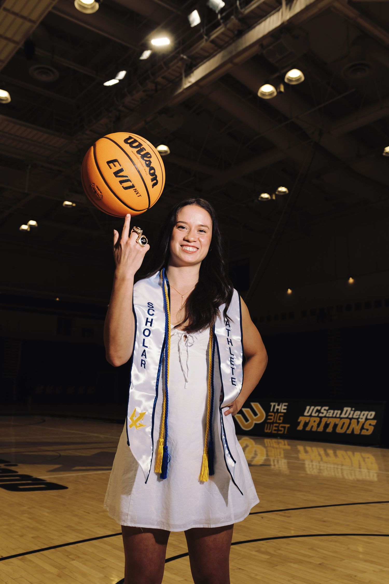 Female graduate in white dress spins basketball on finger at UC San Diego Tritons arena.