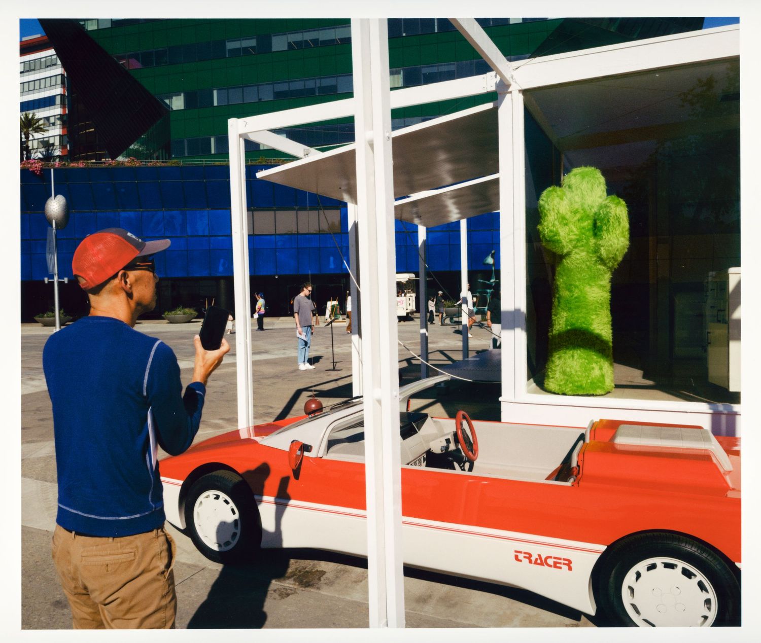 Man in red cap photographs a green cactus sculpture near a vintage red and white car outdoors.