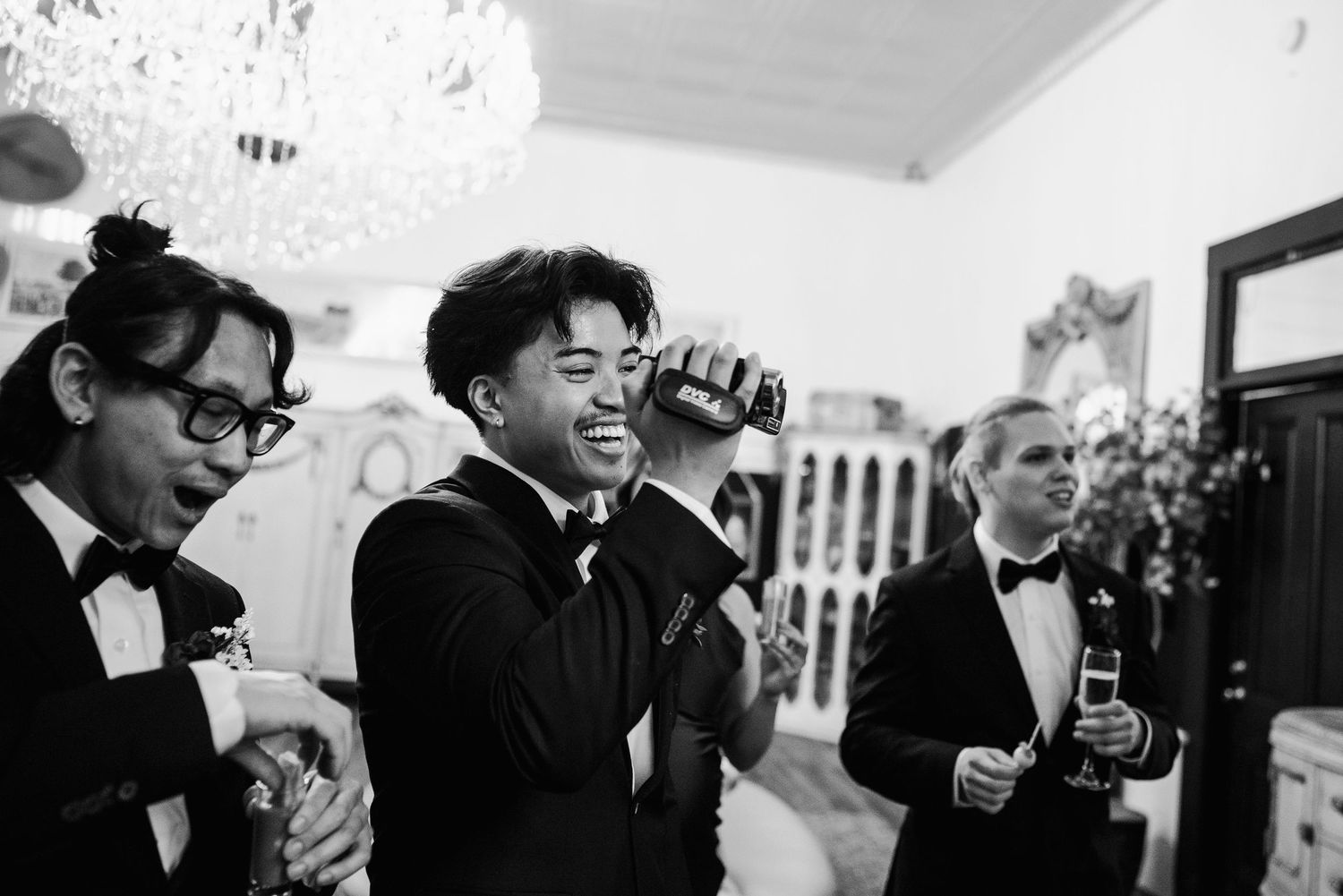Groom laughs while drinking from bottle with groomsmen in tuxedos at black and white wedding reception.