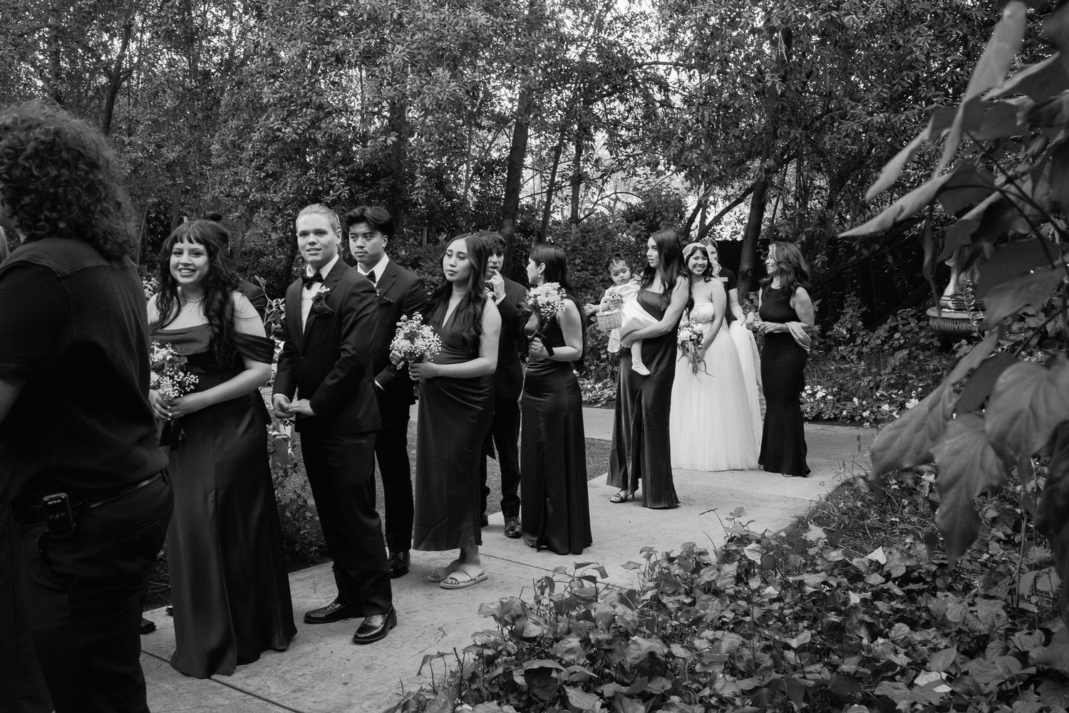 Black and white photo of wedding party gathered outdoors in garden setting.