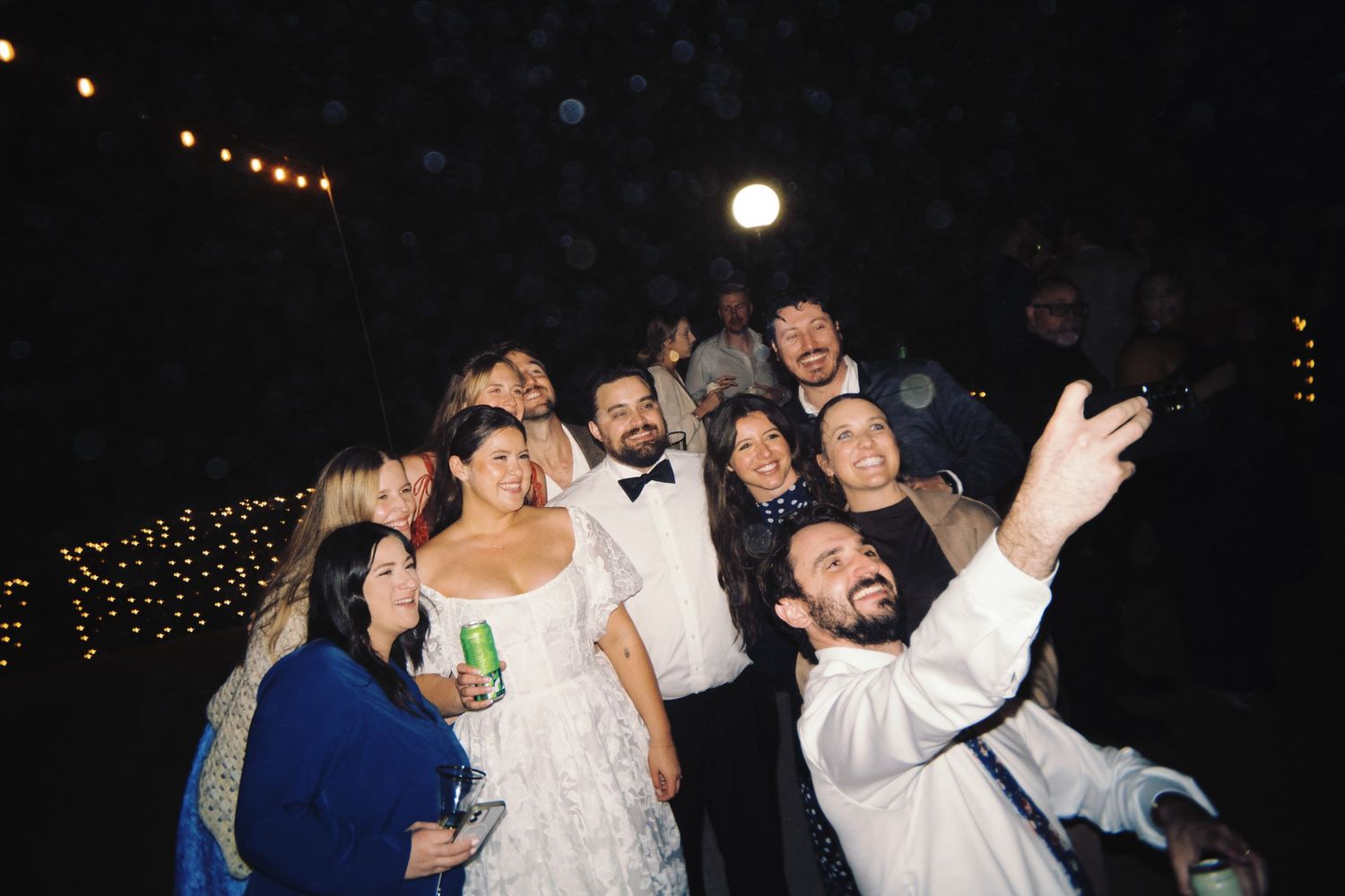 Wedding group selfie at night with bride in white dress and groom in bow tie surrounded by smiling guests under string lights.