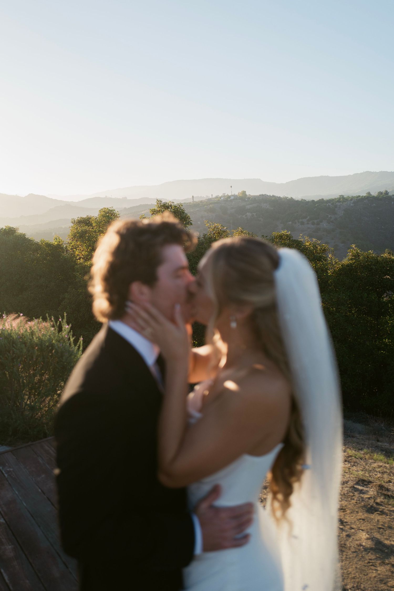Newlyweds share a blurred romantic kiss at sunset with rolling hills and mountains in the background.