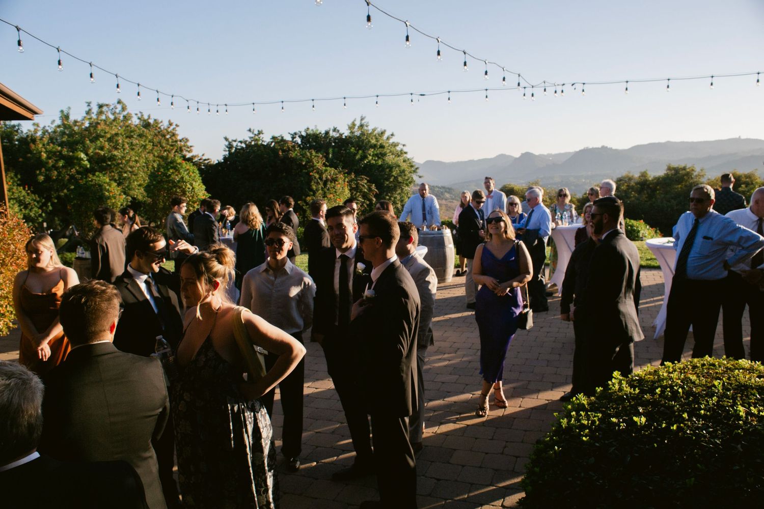 Wedding guests mingling on a patio with string lights overhead and stunning mountain views in warm sunlight.