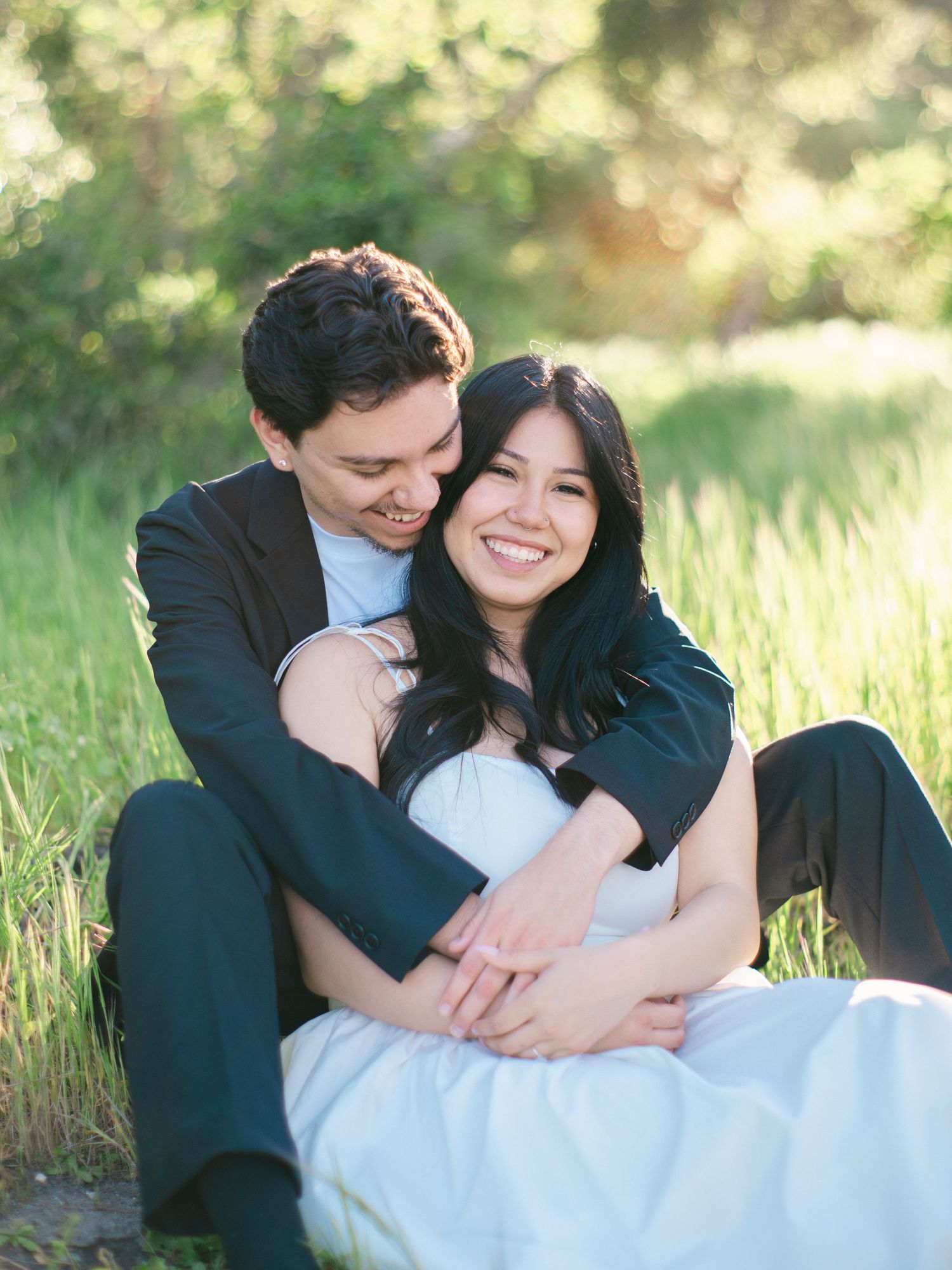 Happy couple sits together in green grass, man embracing woman from behind during engagement photos.