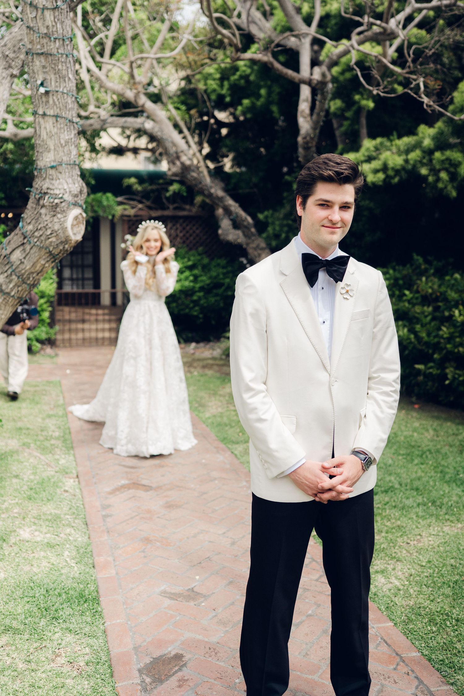 Groom in white tuxedo stands on garden path as bride in lace gown approaches for first look.
