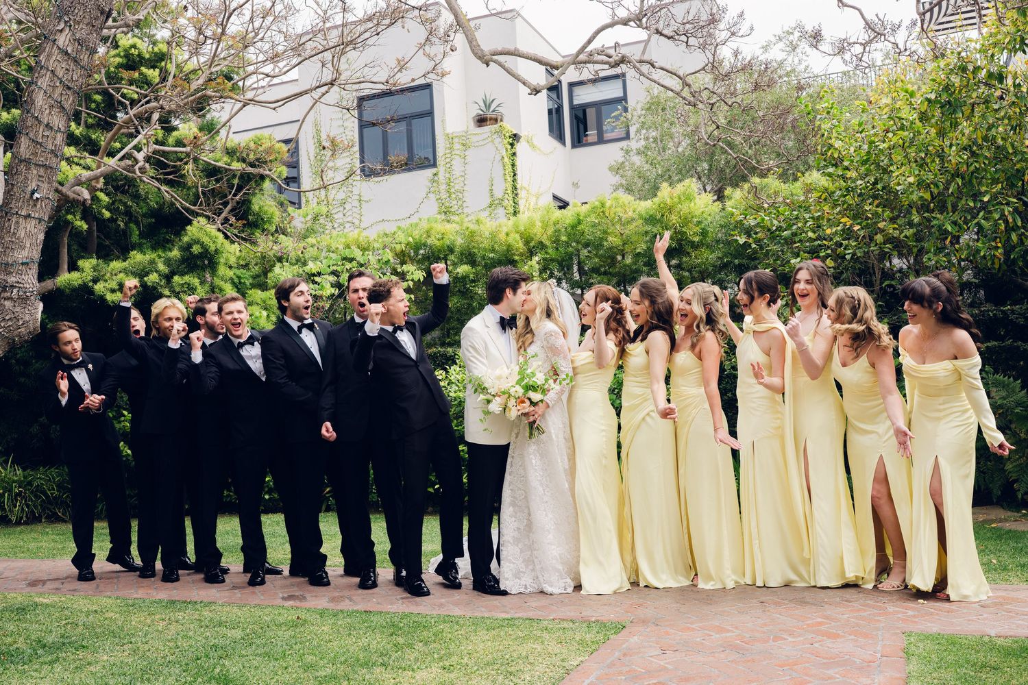 Wedding party in yellow bridesmaid dresses and black tuxedos celebrate outdoors in garden.