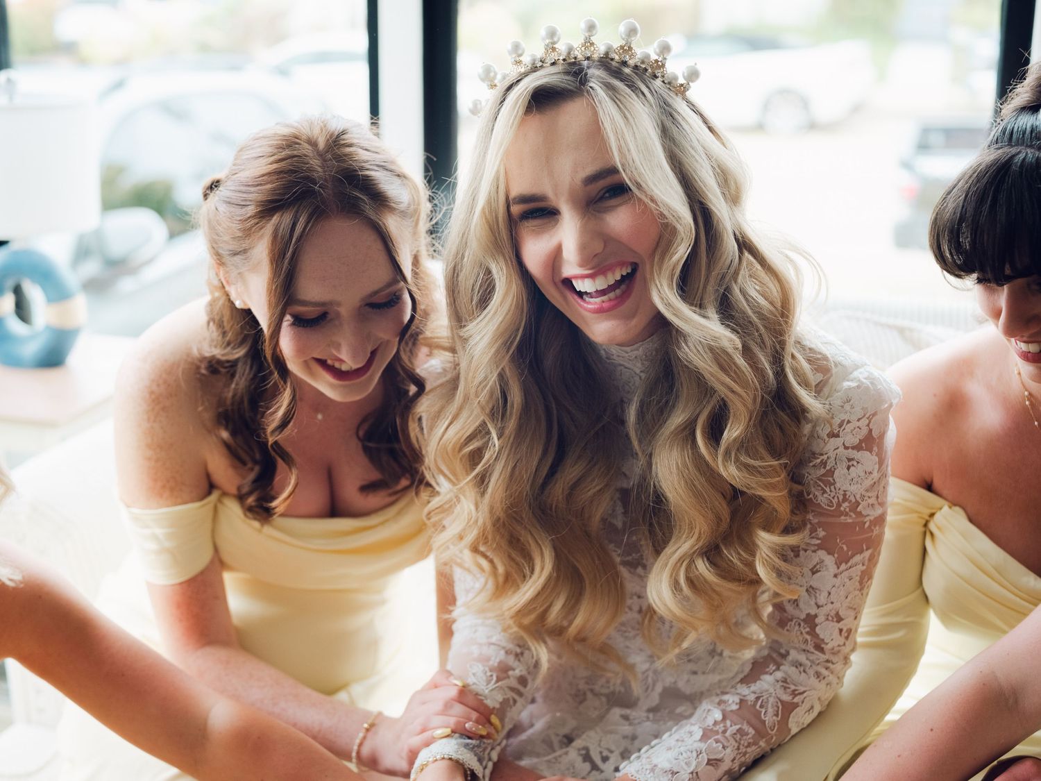 Bride wearing lace gown and tiara laughs joyfully with bridesmaids in yellow off-shoulder dresses during wedding preparations.