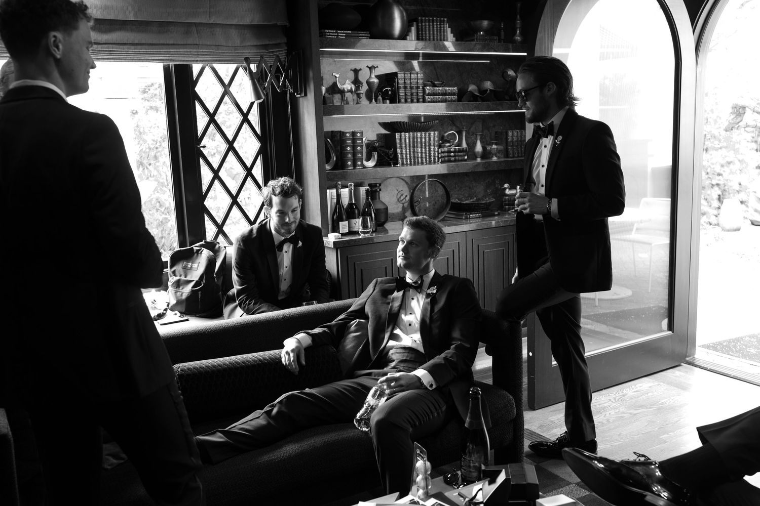Black and white photo of groomsmen in tuxedos relaxing in an elegant room before a wedding ceremony.