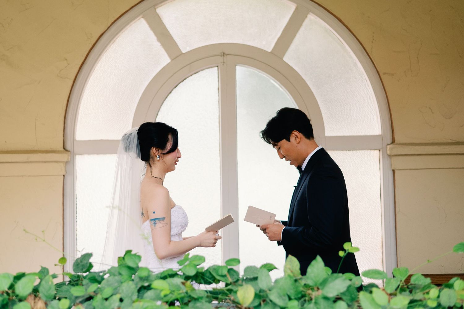Bride and groom exchanging private vows by arched window on wedding day.