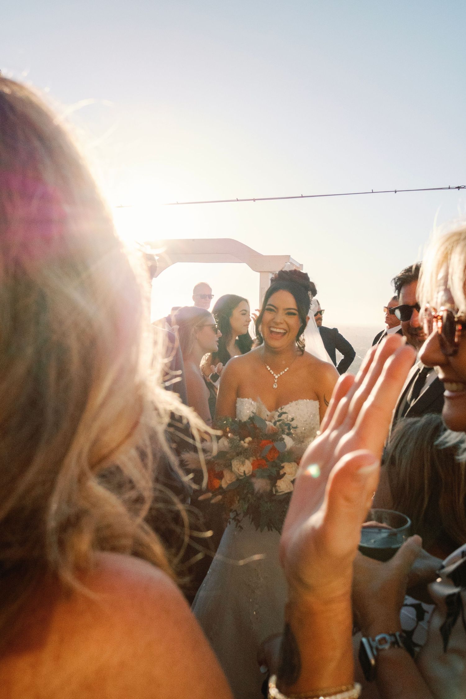 Radiant bride in strapless gown holding bouquet walks down aisle at golden hour outdoor wedding ceremony.